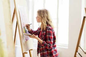 art school, creativity and people concept - student girl or artist with easel, palette and paint brush painting still life picture at studio